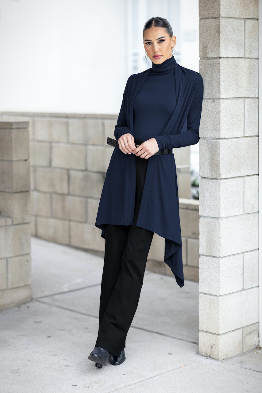 A woman wearing a long-sleeve navy blue wrap cardigan over a matching sleeveless turtleneck top, paired with black pants and black shoes. Leaning elegantly against a cement wall.