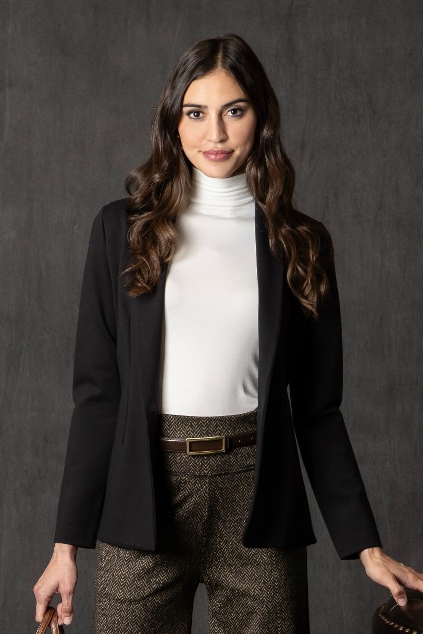 Woman wearing a black blazer, white mock neck, and brown herringbone patterned pants against a dark background