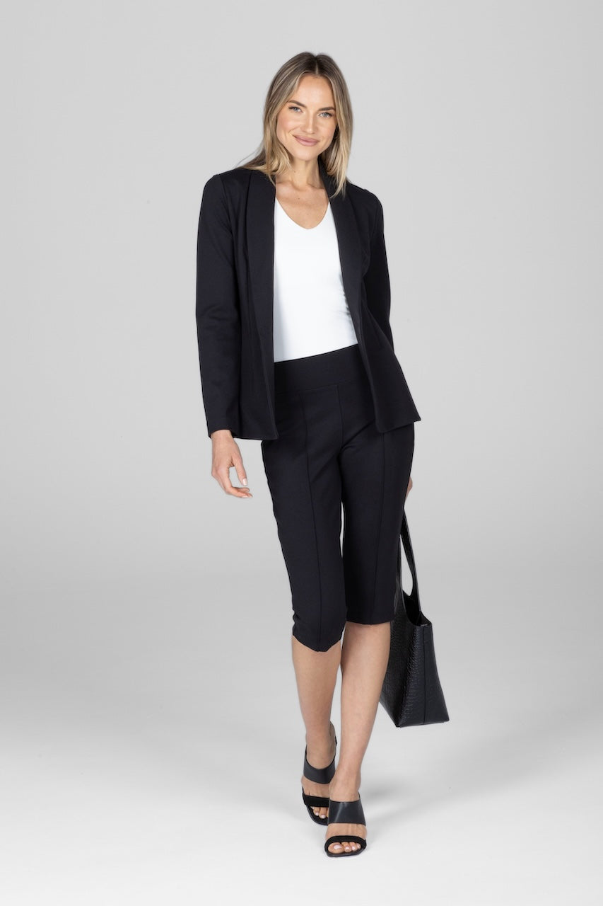 A woman standing against a white background wearing a black unbuttoned blazer, white v-neck tank top, black knee pants, and black shoes. She is holding a black bag.
