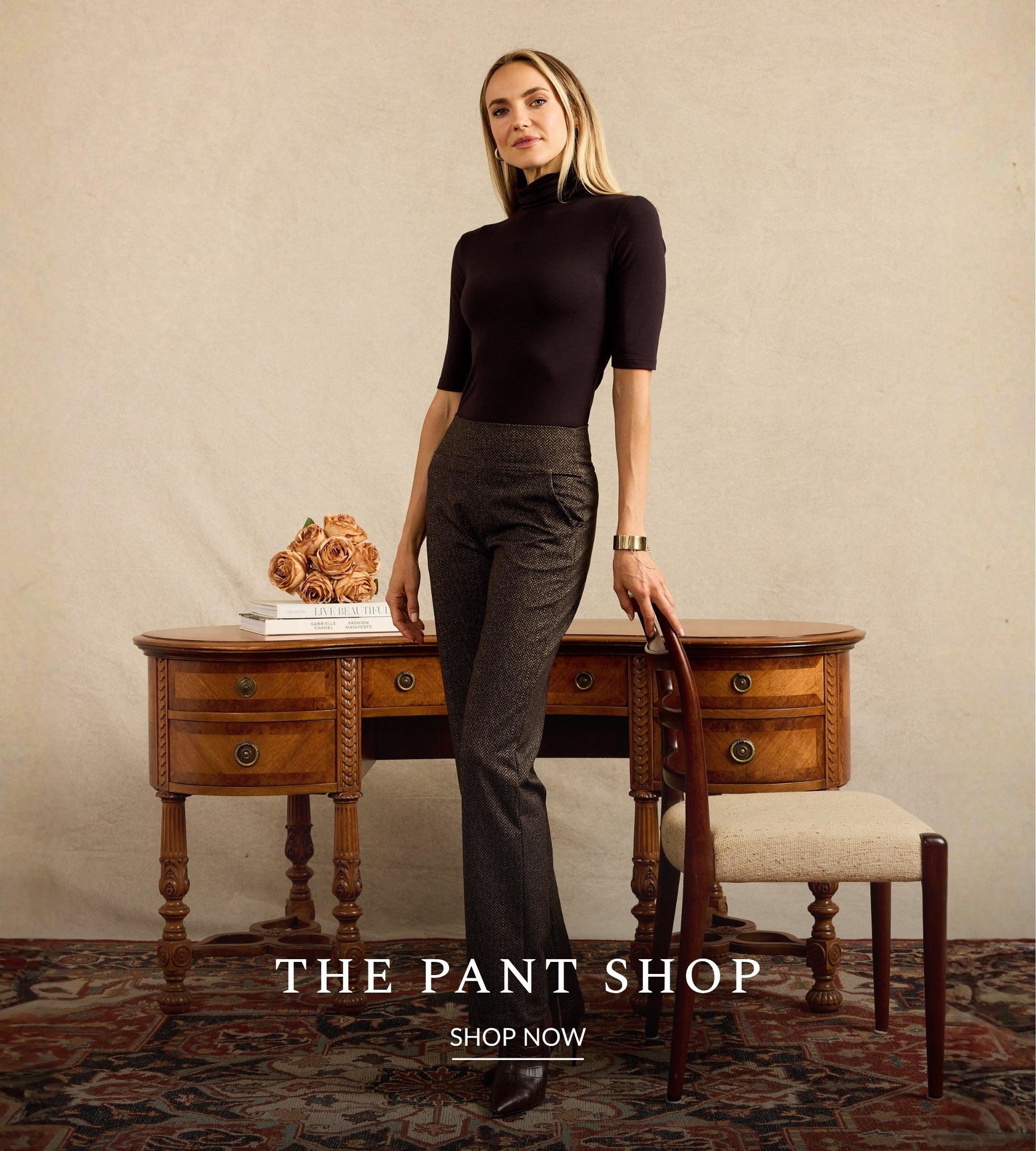 A woman stands in a styled room wearing a fitted brown short-sleeve mock-neck top and dark brown straight leg trousers, leaning against a wooden desk next to a chair. Text at the bottom reads 'THE PANT SHOP' and 'SHOP NOW'.