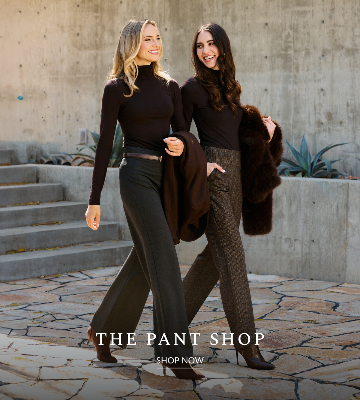 Two women walking outdoors in front of a concrete wall, each wearing a dark brown mock-neck long-sleeve top and different styles of dark pants. One wears black wide-leg pants and the other wears textured taupe-brown straight-leg pants.