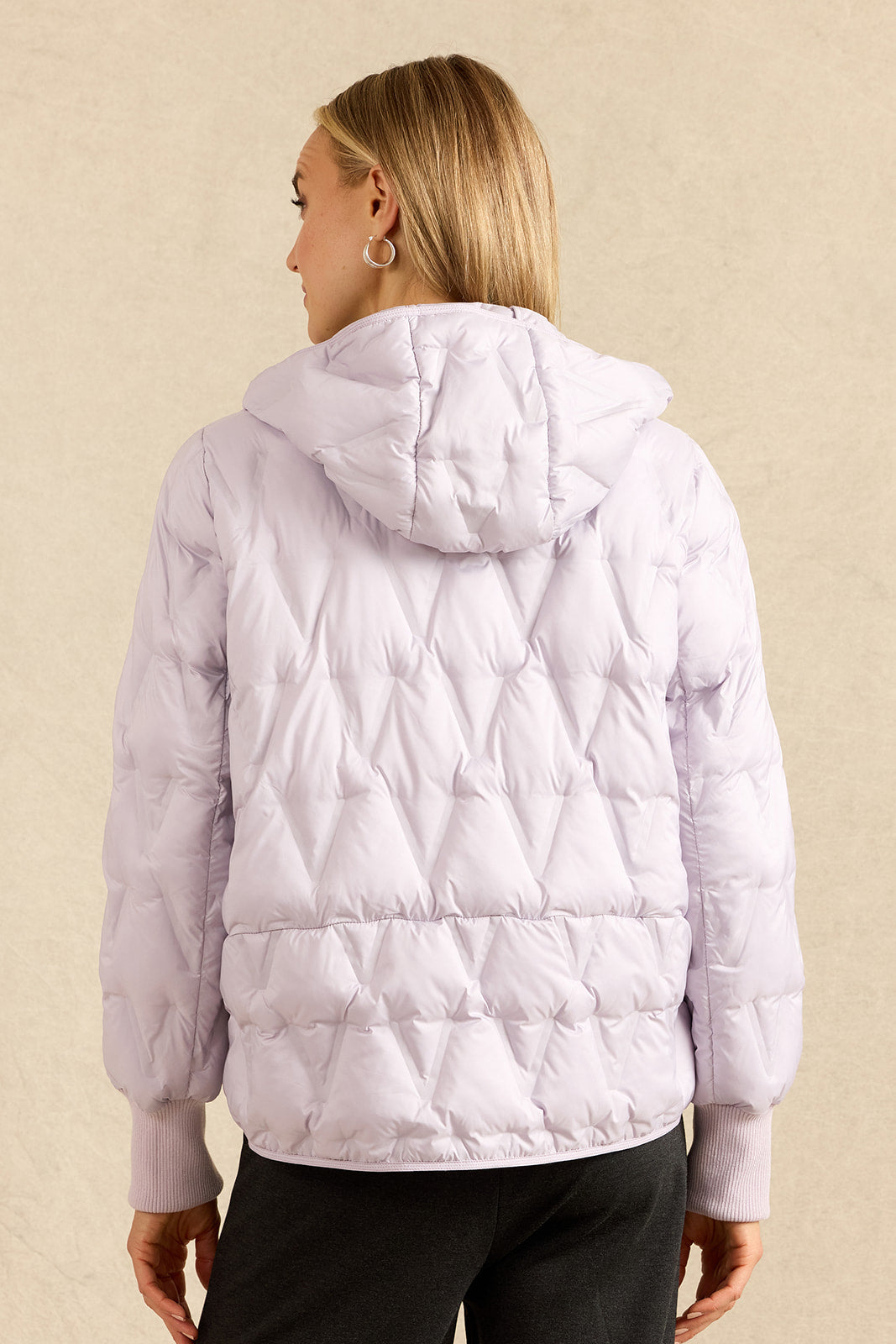 A woman viewed from the back wearing a light lavender quilted puffer jacket with a hood, paired with black pants, against a textured beige background.