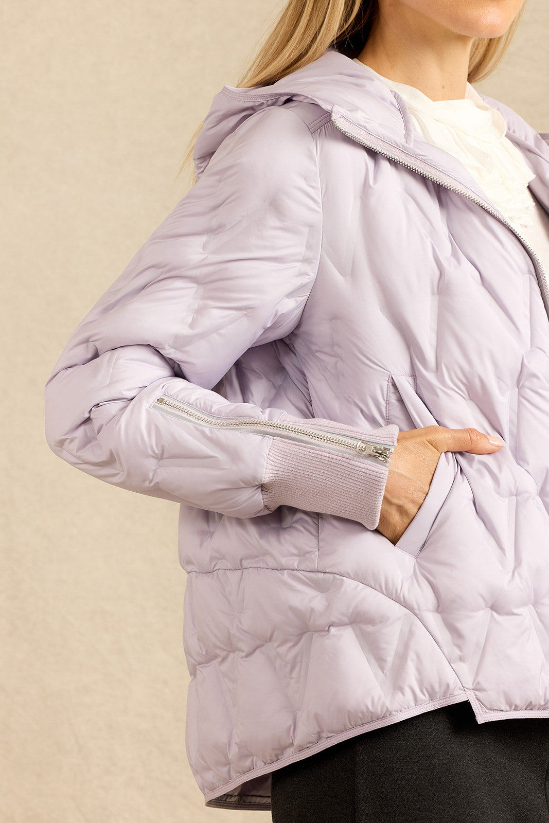 A close-up of a woman wearing a light lavender puffer jacket with a hood, zippered cuffs, and side pockets, paired with a white top and dark pants against a neutral beige background.
