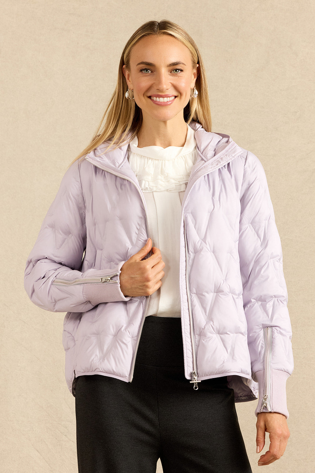 A woman modeling a light lavender quilted puffer jacket with a hood and silver zipper details, worn over a white top and charcoal pants, against a textured beige background.