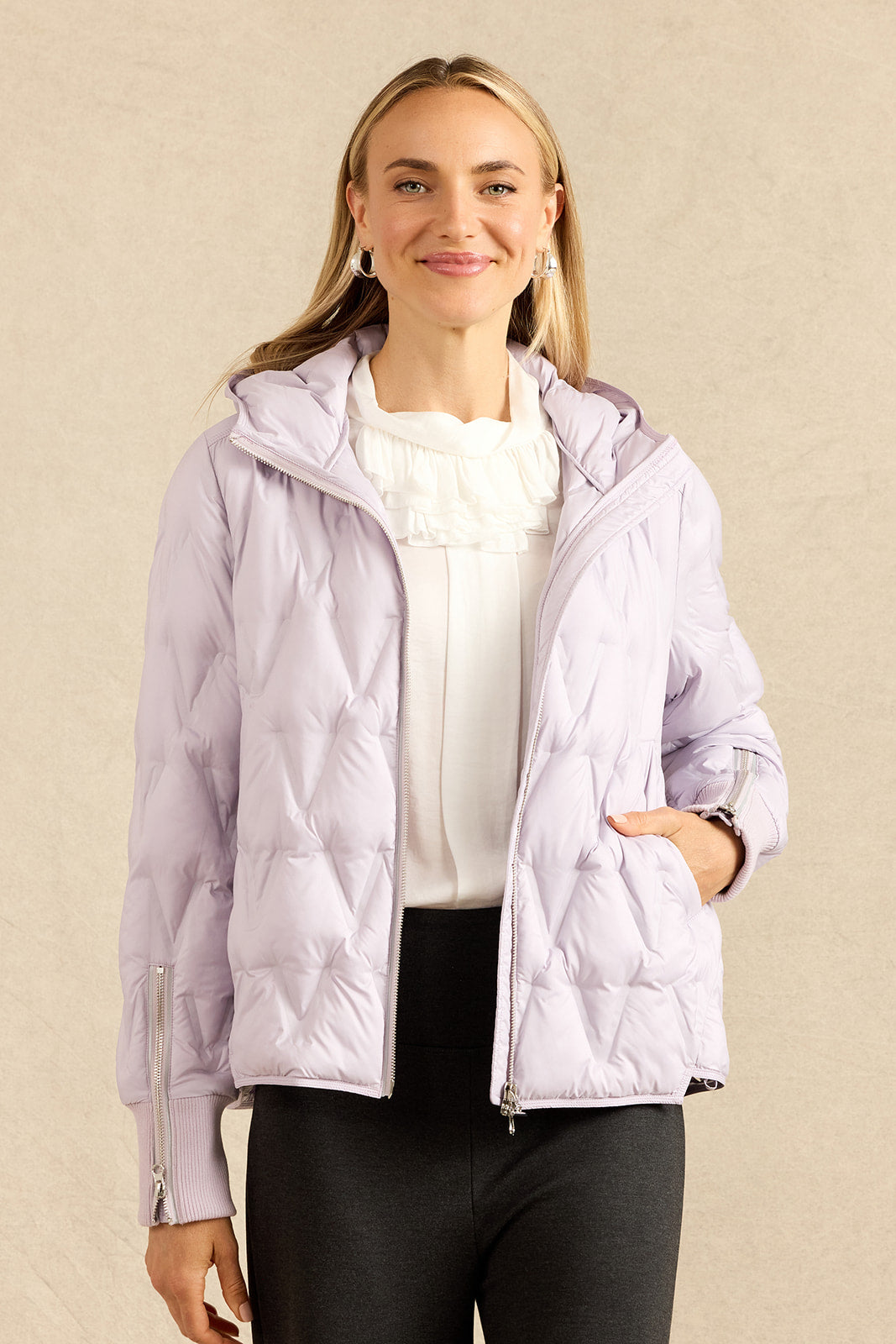 A woman modeling a light lavender hooded puffer jacket with silver zipper details, worn over a white ruffled blouse and black pants, against a textured beige background.