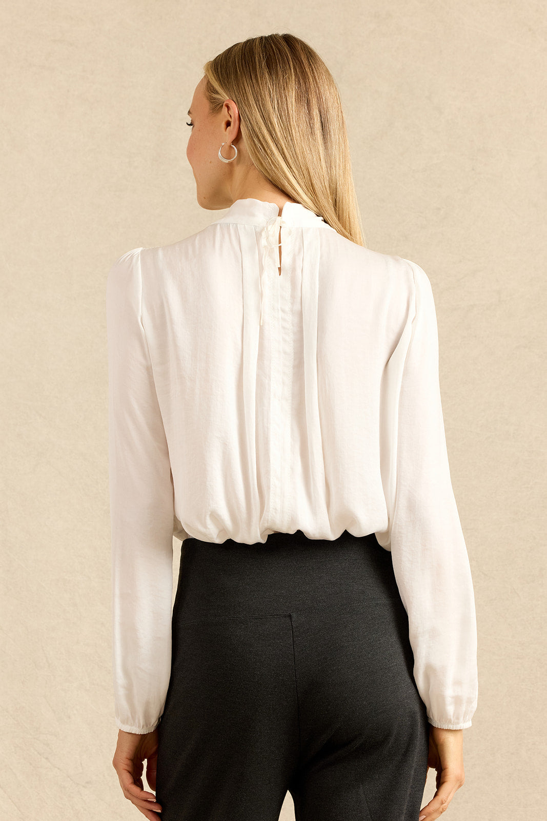 A woman shown from the back wearing a white long-sleeve blouse with gathered pleating at the back and charcoal pants, standing against a light beige textured wall.