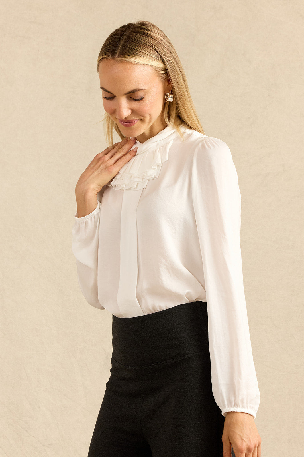 A woman wearing a white long-sleeve blouse with a ruffled high neck and black trousers, standing against a textured beige background.