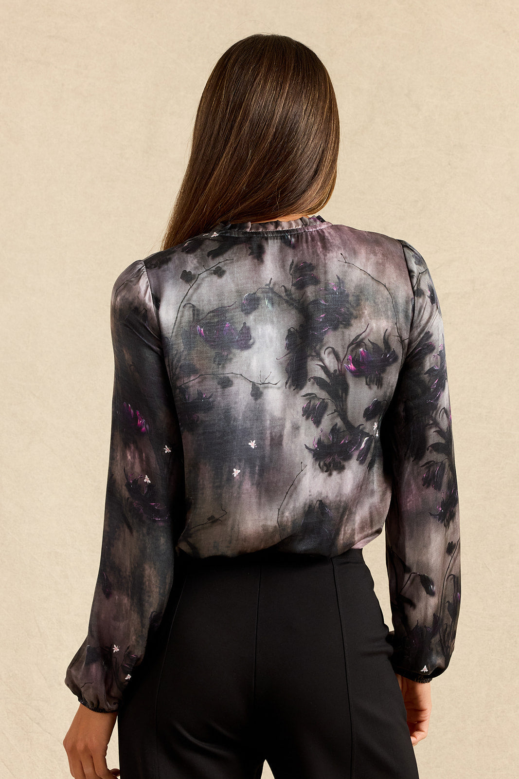 A woman viewed from the back wearing a sheer long-sleeve blouse with a dark floral and abstract print in black, gray, and purple, paired with black pants against a textured beige background.
