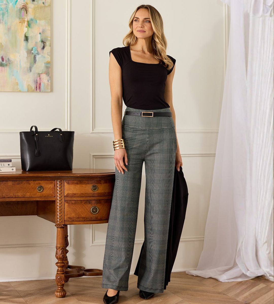Woman in square neck black top and black and white plaid pants standing in a room with a wooden desk and black handbag.
