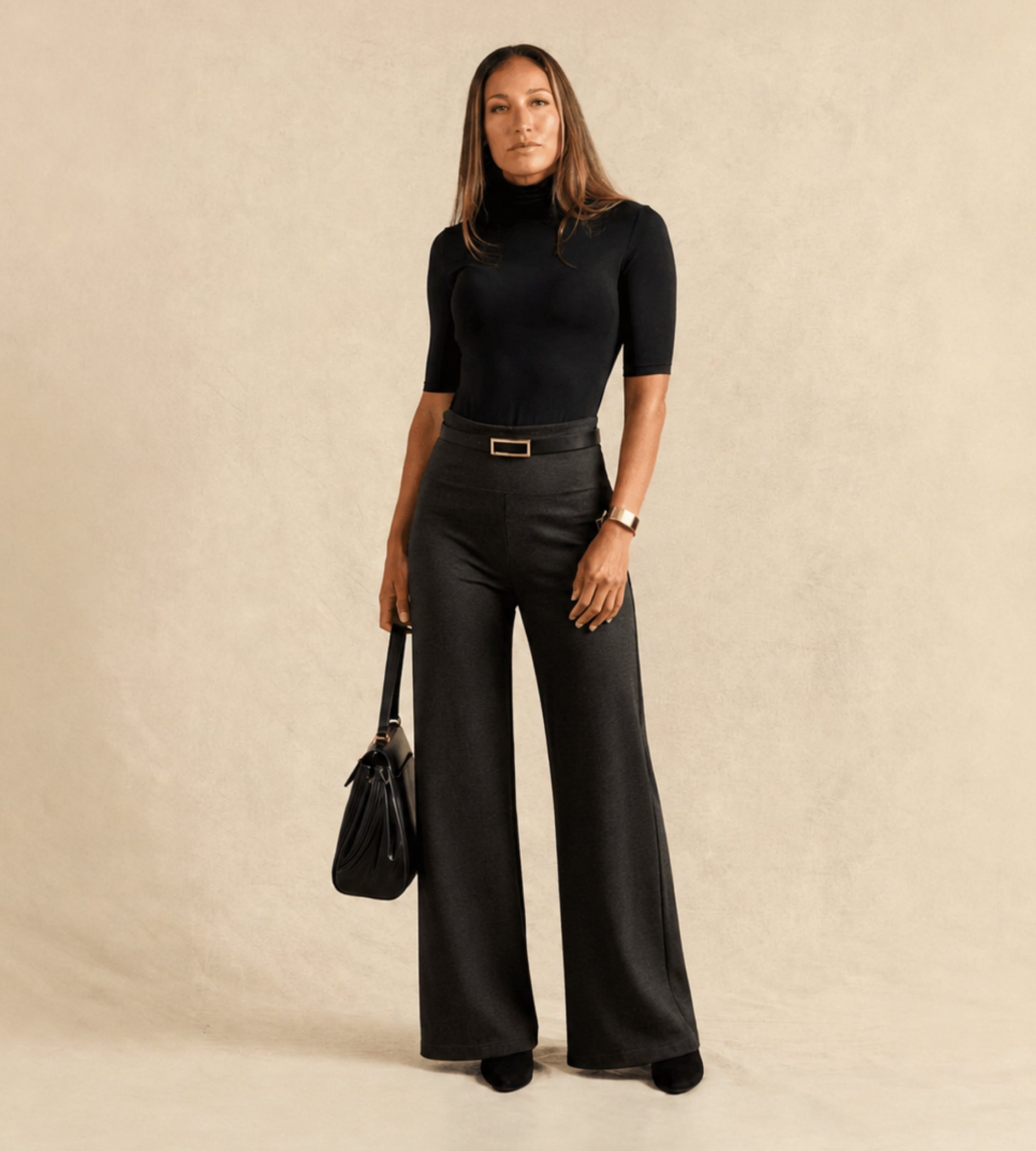 A woman stands against a warm beige textured backdrop wearing a black short-sleeve mock-neck top, black wide-leg trousers with a thin belt, and holding a black handbag.