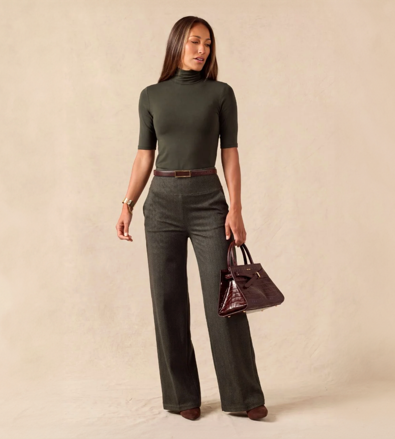 A woman models a dark olive green mock-neck short-sleeve top paired with matching wide-leg trousers, a brown belt, and a dark brown handbag, set against a plain beige studio backdrop.