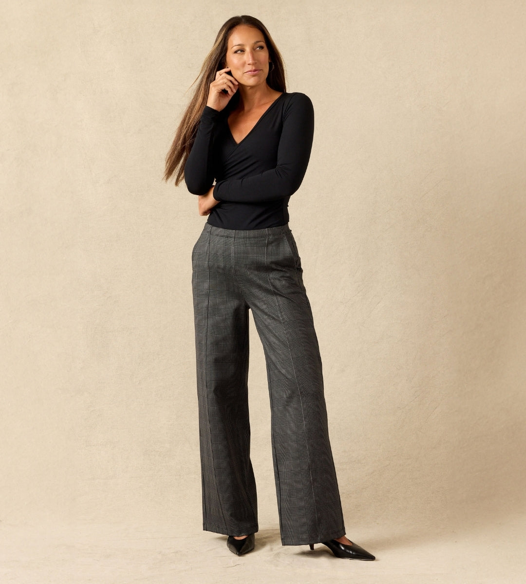 A woman modeling a black long-sleeve V-neck top and dark gray wide-leg plaid trousers, standing against a textured beige studio backdrop.