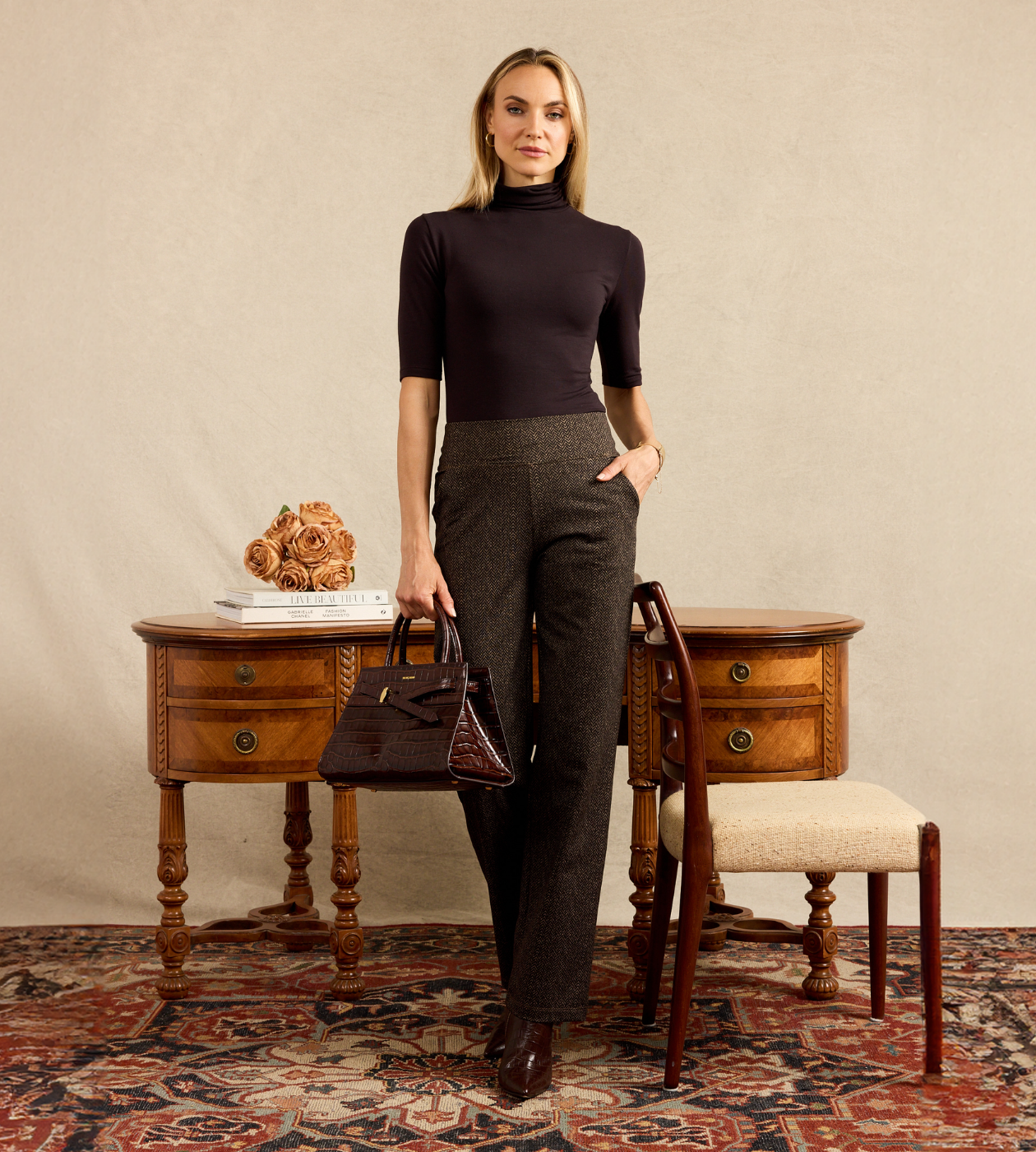 A woman stands in a styled room wearing a dark brown mock-neck short-sleeve top and matching dark brown textured trousers, holding a dark brown handbag. She is positioned in front of a vintage wooden desk and chair against a textured beige wall.