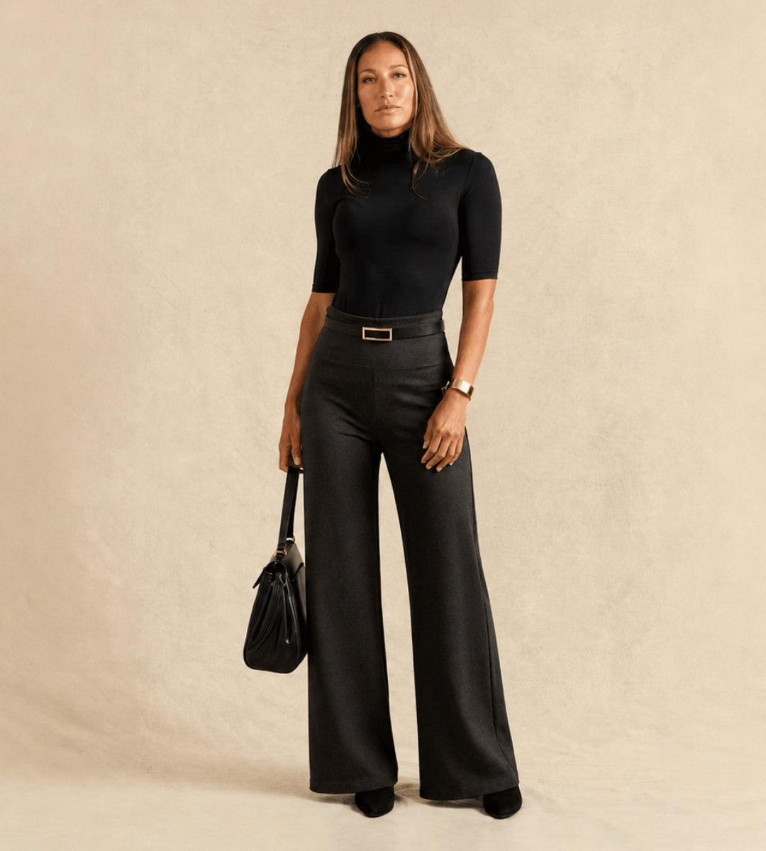 A woman stands against a warm beige textured backdrop wearing a fitted black short-sleeve mock-neck top and charcoal wide-leg trousers with a thin belt. She is holding a black handbag.