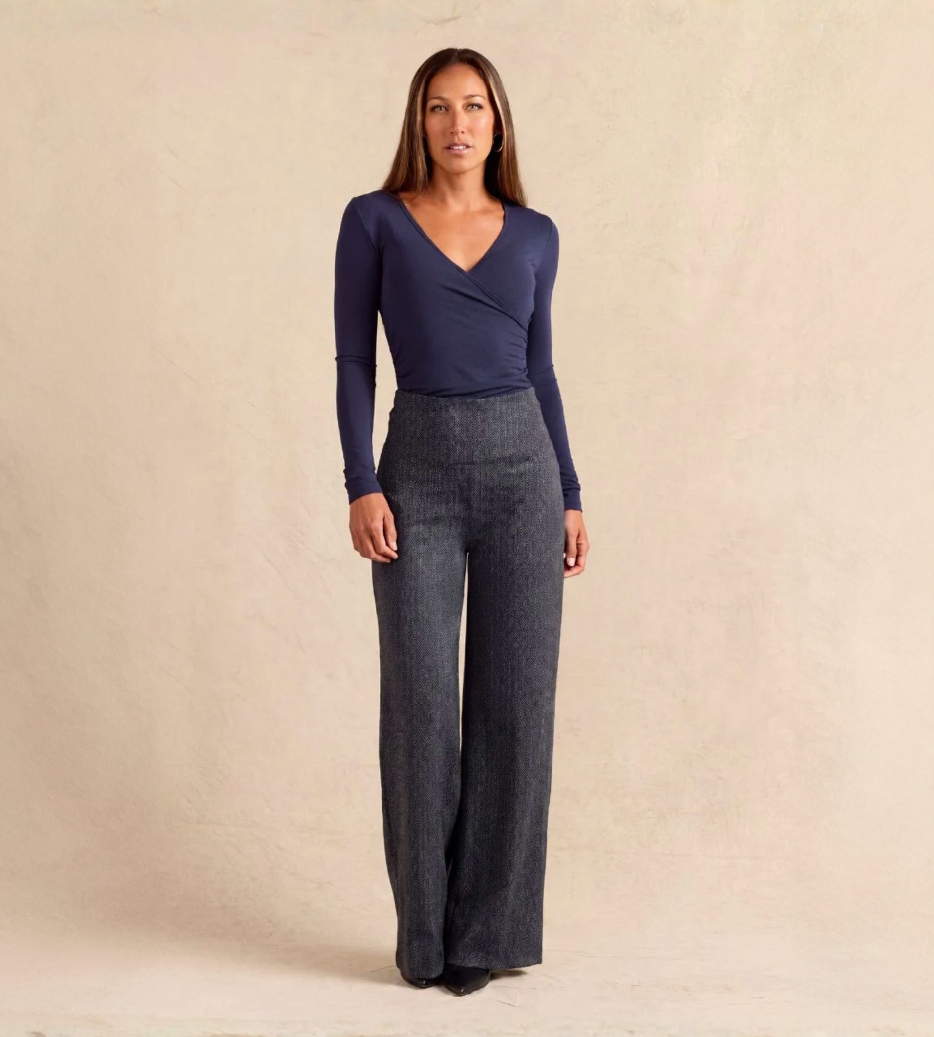 A woman modeling a navy blue long-sleeve wrap top and dark gray wide-leg trousers against a textured beige studio backdrop.