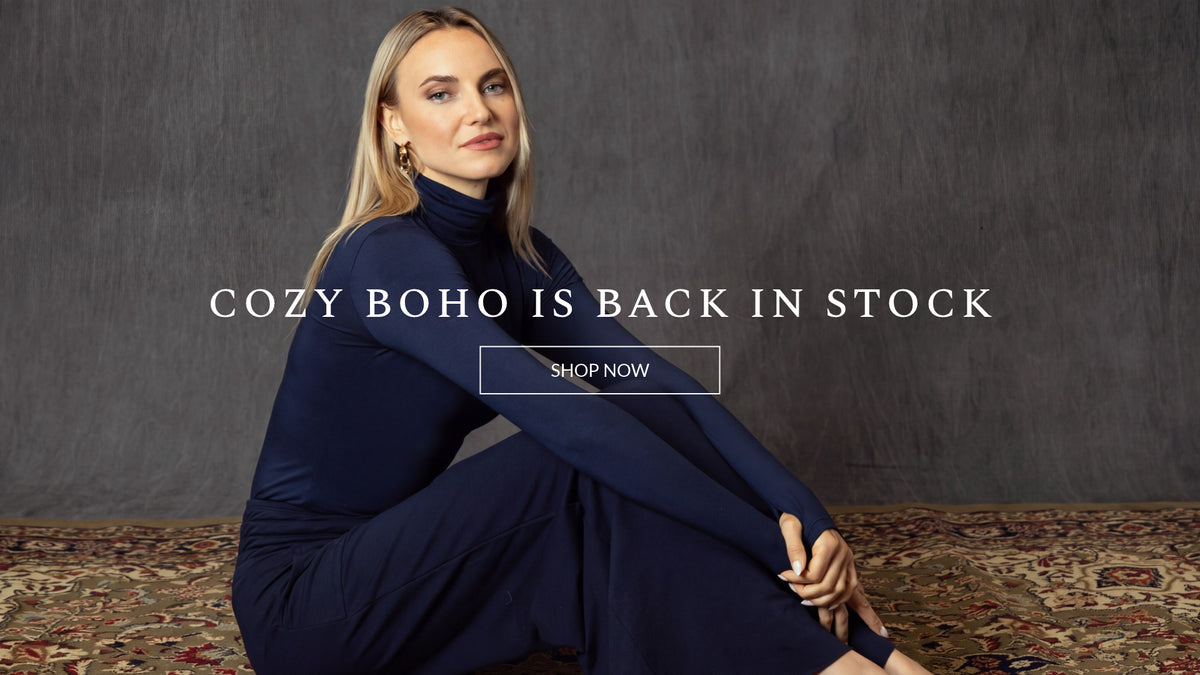 A woman sits cross-legged on a patterned rug against a dark textured wall, wearing a navy blue mock-neck long-sleeve dress. White text overlay reads 'COZY BOHO IS BACK IN STOCK' with a 'SHOP NOW' button.