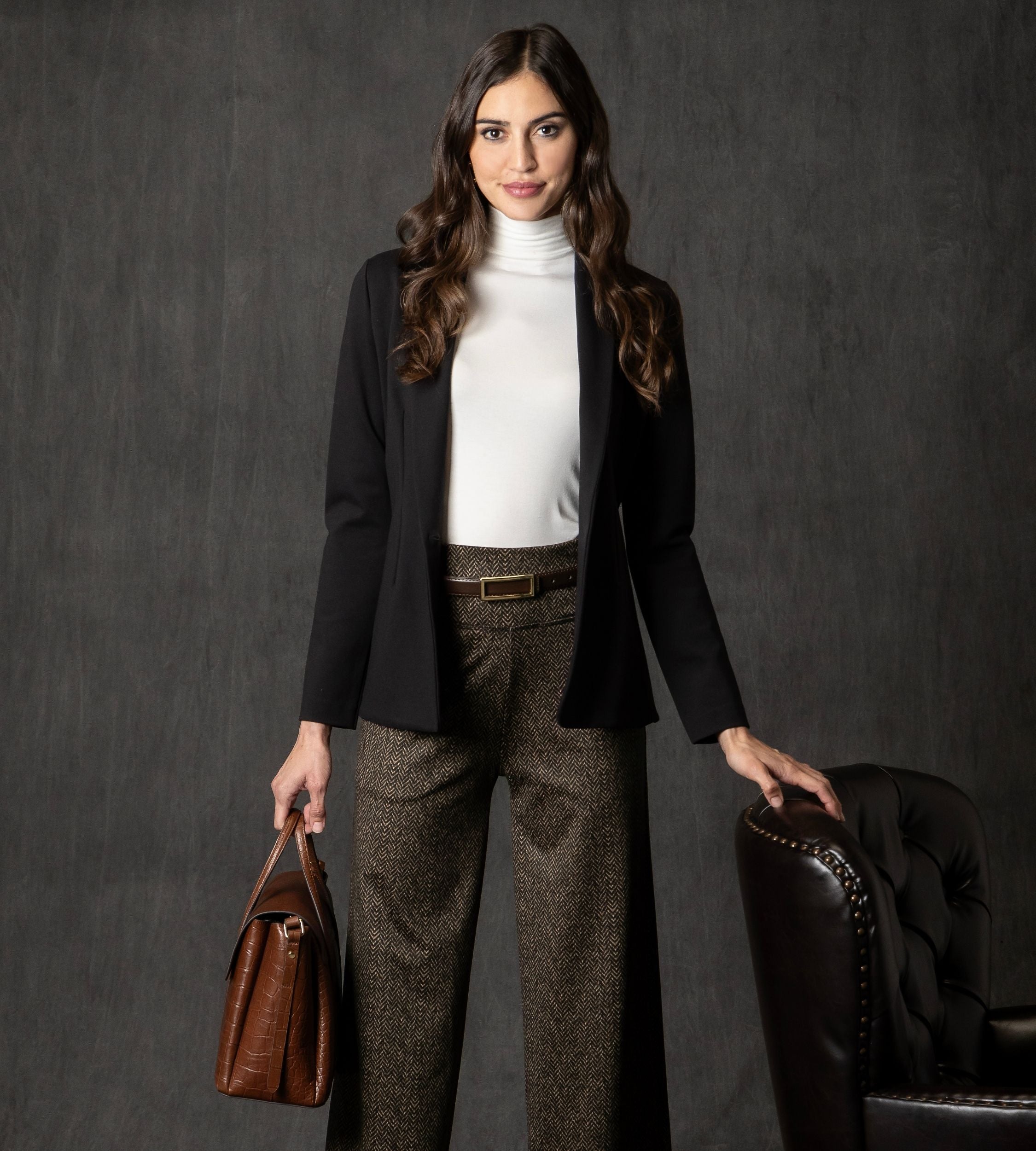 Woman in professional blazer and belt with herringbone camel high waisted pants holding a brown leather bag against a dark background