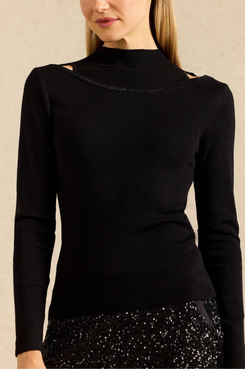 A woman wearing a fitted black long-sleeve mock-neck top with small cutout details at the shoulders and a subtle shimmering trim along the neckline, paired with a black sequined skirt against a neutral beige background.