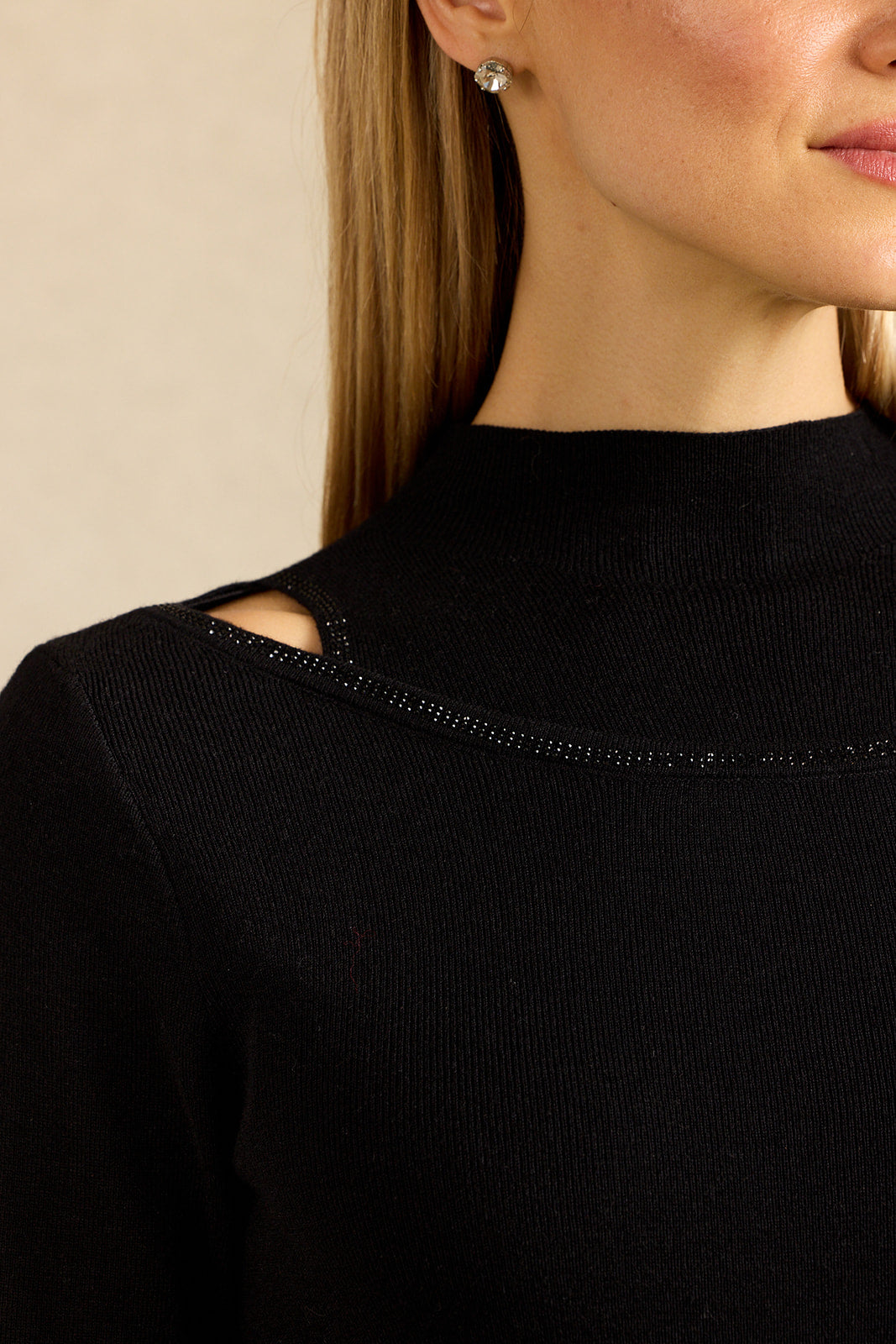 Close-up of a woman wearing a black ribbed mock neck top with a shoulder cutout and a line of small rhinestones around the neckline. She is wearing a small silver stud earring against a plain beige background.