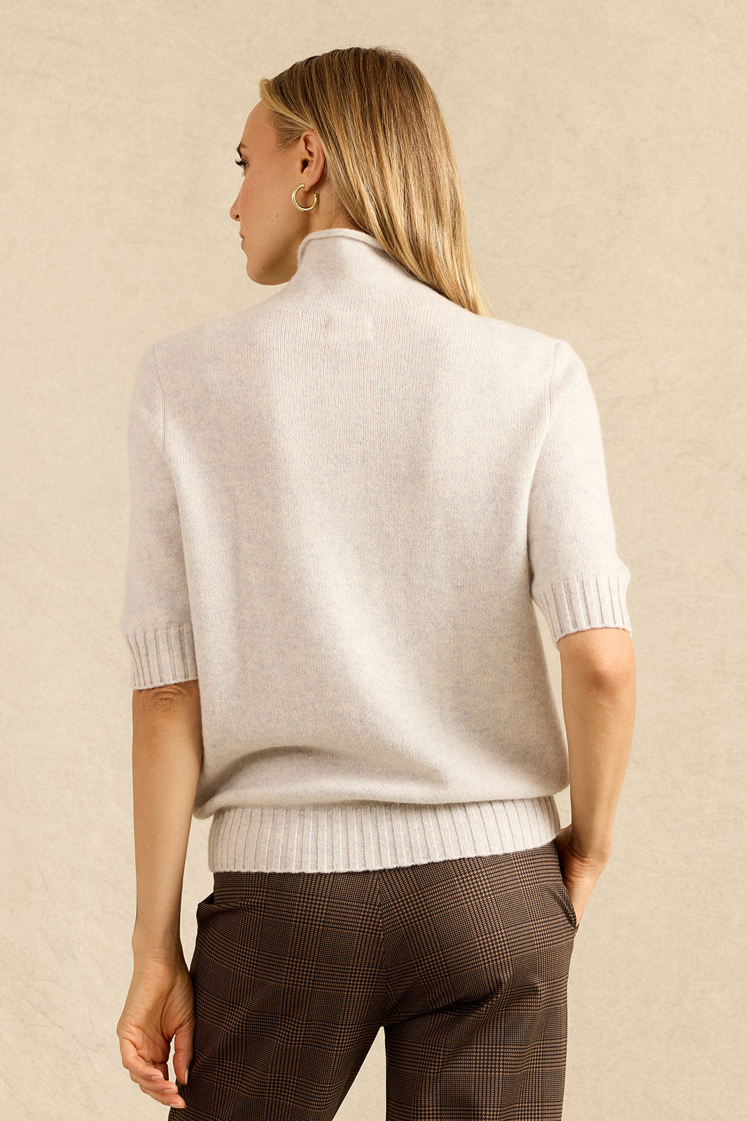 A woman shown from the back wearing a light cream knit mock-neck sweater with short sleeves and ribbed cuffs and hem, paired with brown plaid trousers, against a textured beige background.