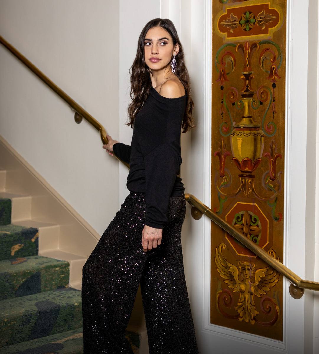 Woman in black off-shoulder sweater and black sequin wide leg pants standing on a staircase with decorative wall behind.