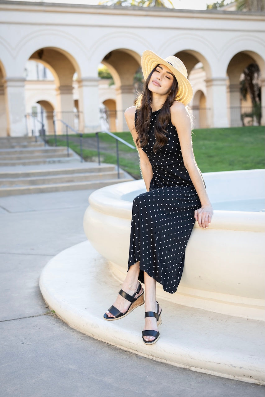 Woman sitting on the edge of a water fountain with eyes closed and wearing a black sleeveless dress with a white dot pattern. Also wearing a beige straw hat.