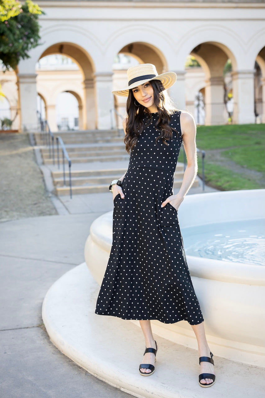 Woman standing in front of a water fountain wearing a sleeveless black dress with white dot pattern as well as wearing a straw hat with black ribbon.