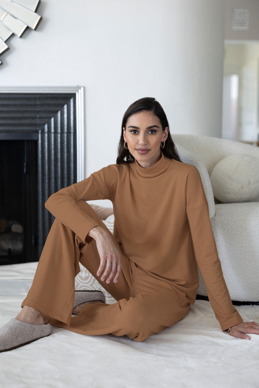 A woman sitting on a cozy surface wearing brown wide-leg camel sweatpants and a matching mockneck long sleeve top, with a decorative fireplace and furniture in the background.