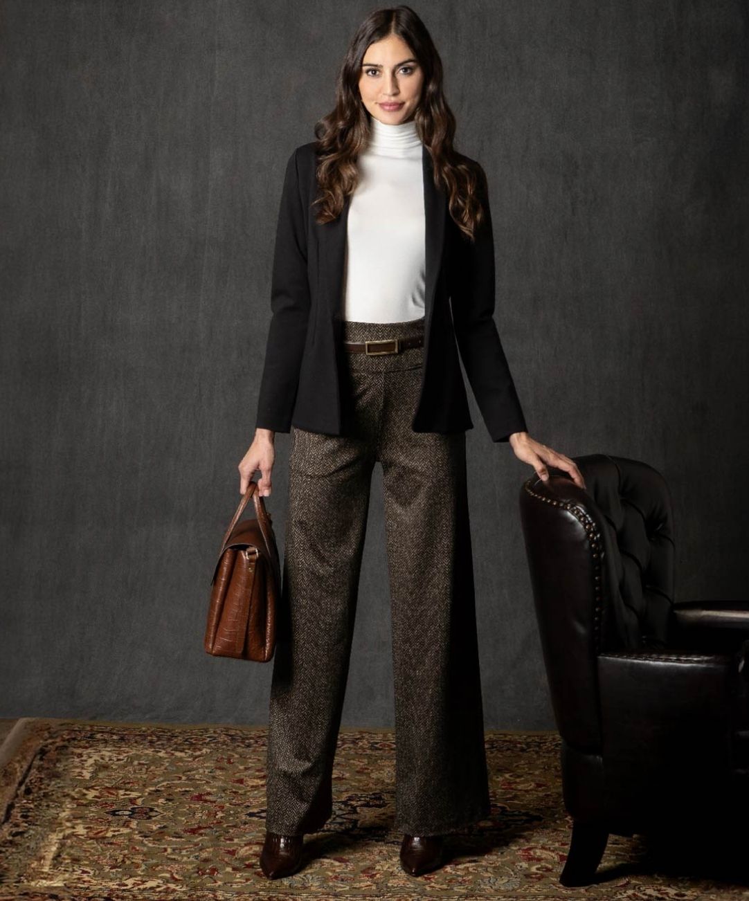 Woman in professional attire with a black blazer, white mockneck and brown trousers, holding a brown leather bag against a dark background.