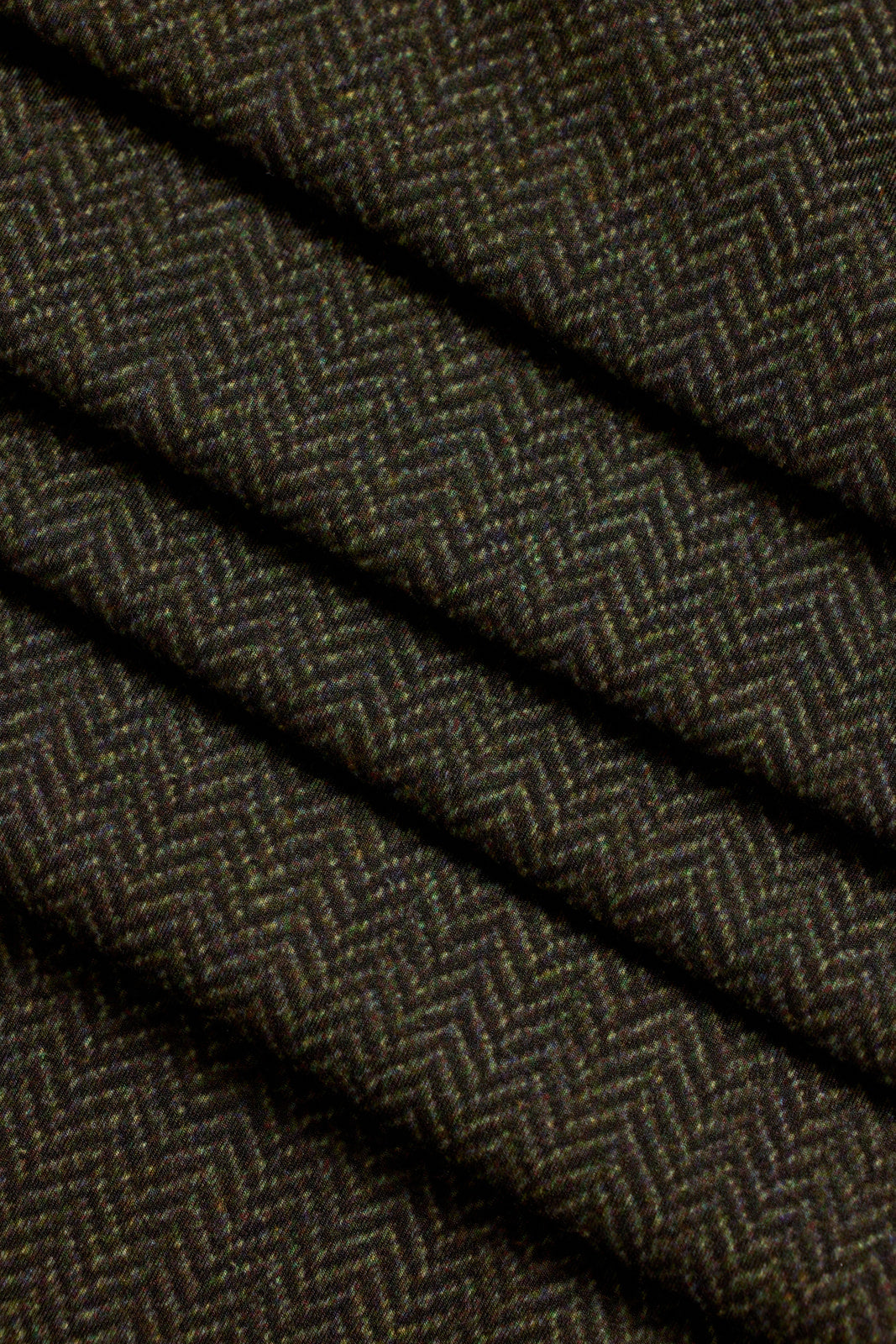 A close-up of dark olive tweed herringbone woven fabric with diagonal folds showing the textile texture.