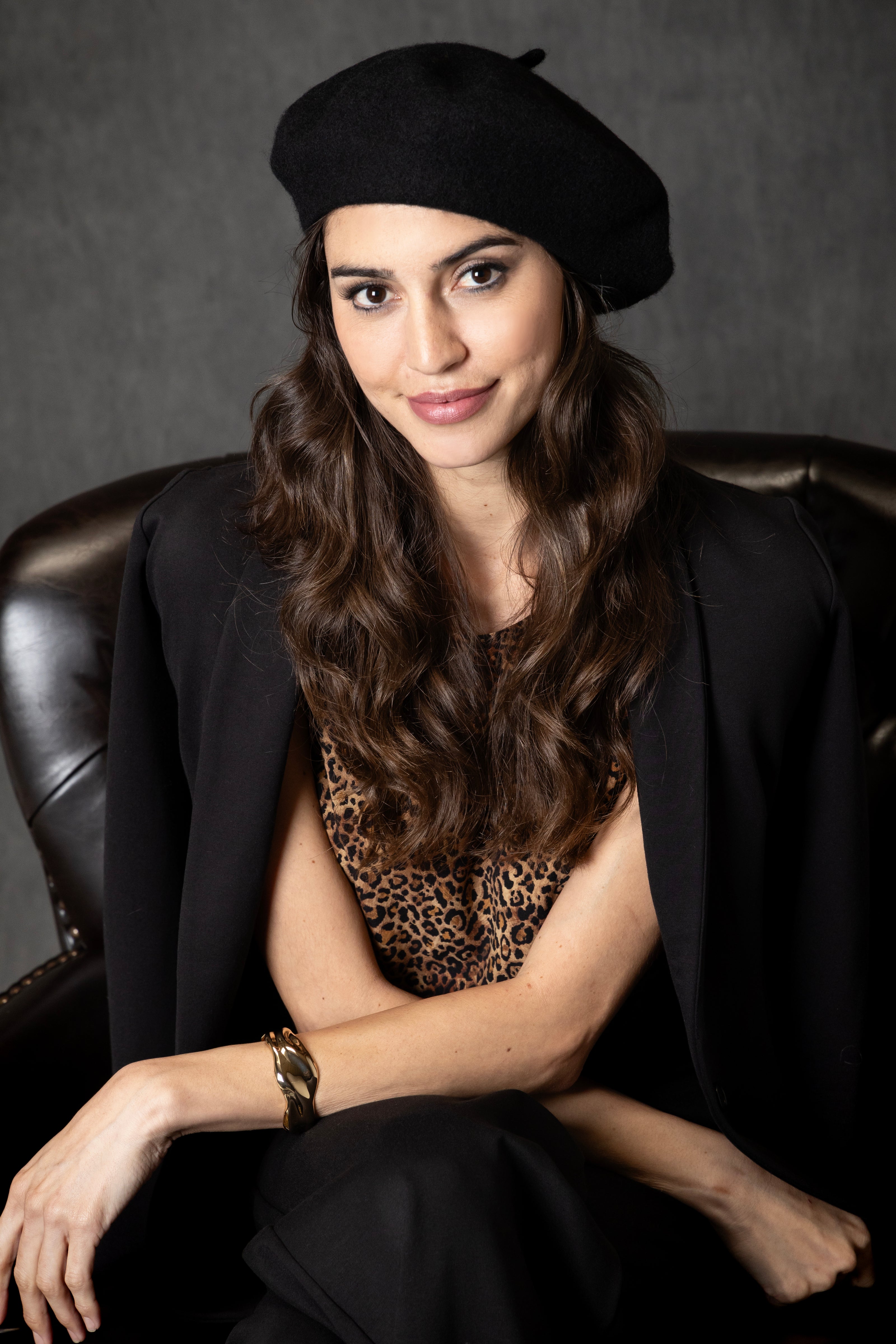Woman wearing a black beret and black blazer with a leopard print top, sitting on a leather chair.