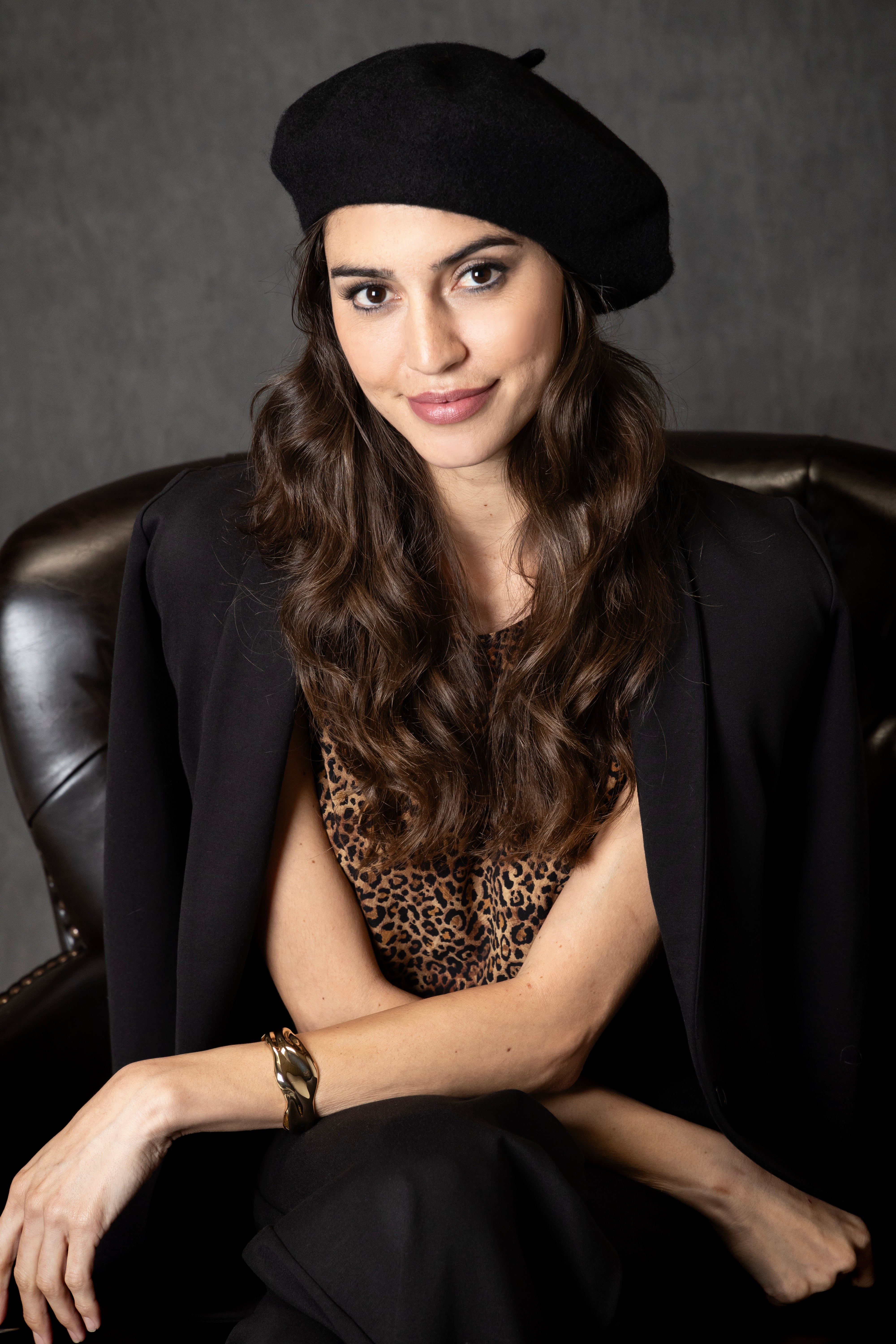 Woman wearing a black beret and black blazer with a leopard print top, sitting on a leather chair.