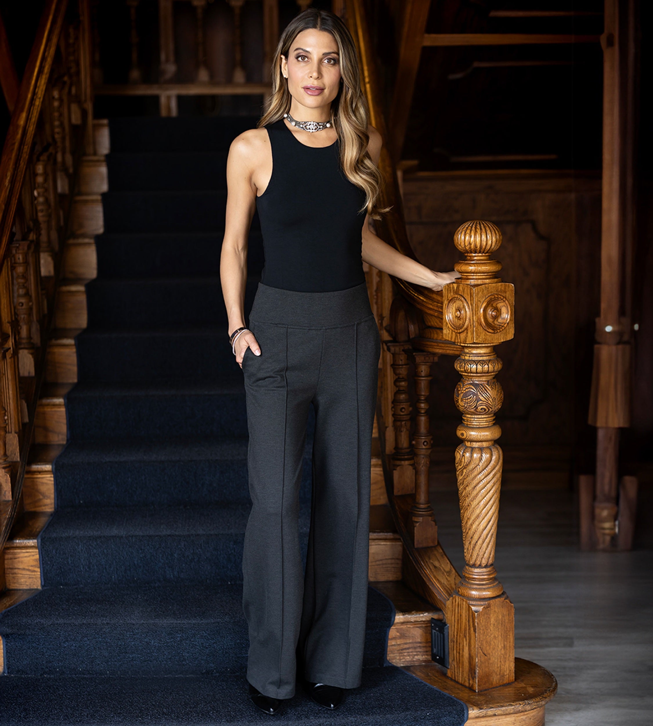 Woman in black sleeveless tank top and gray high waisted pintuck pants standing on a staircase with wooden railings.