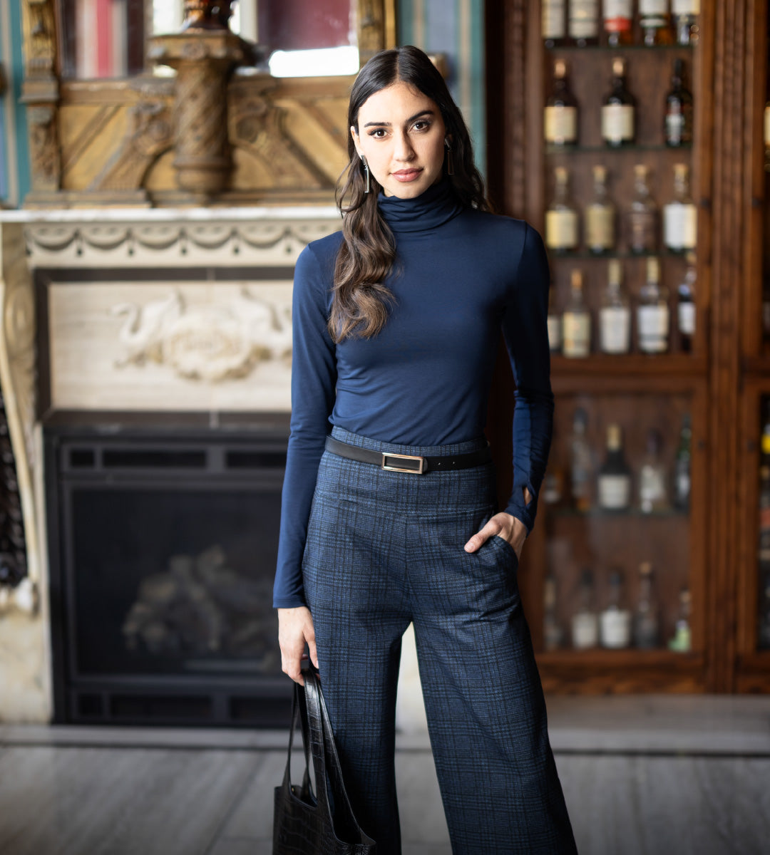 Woman in a blue turtleneck and plaid navy pants standing in a room with a fireplace and shelves.