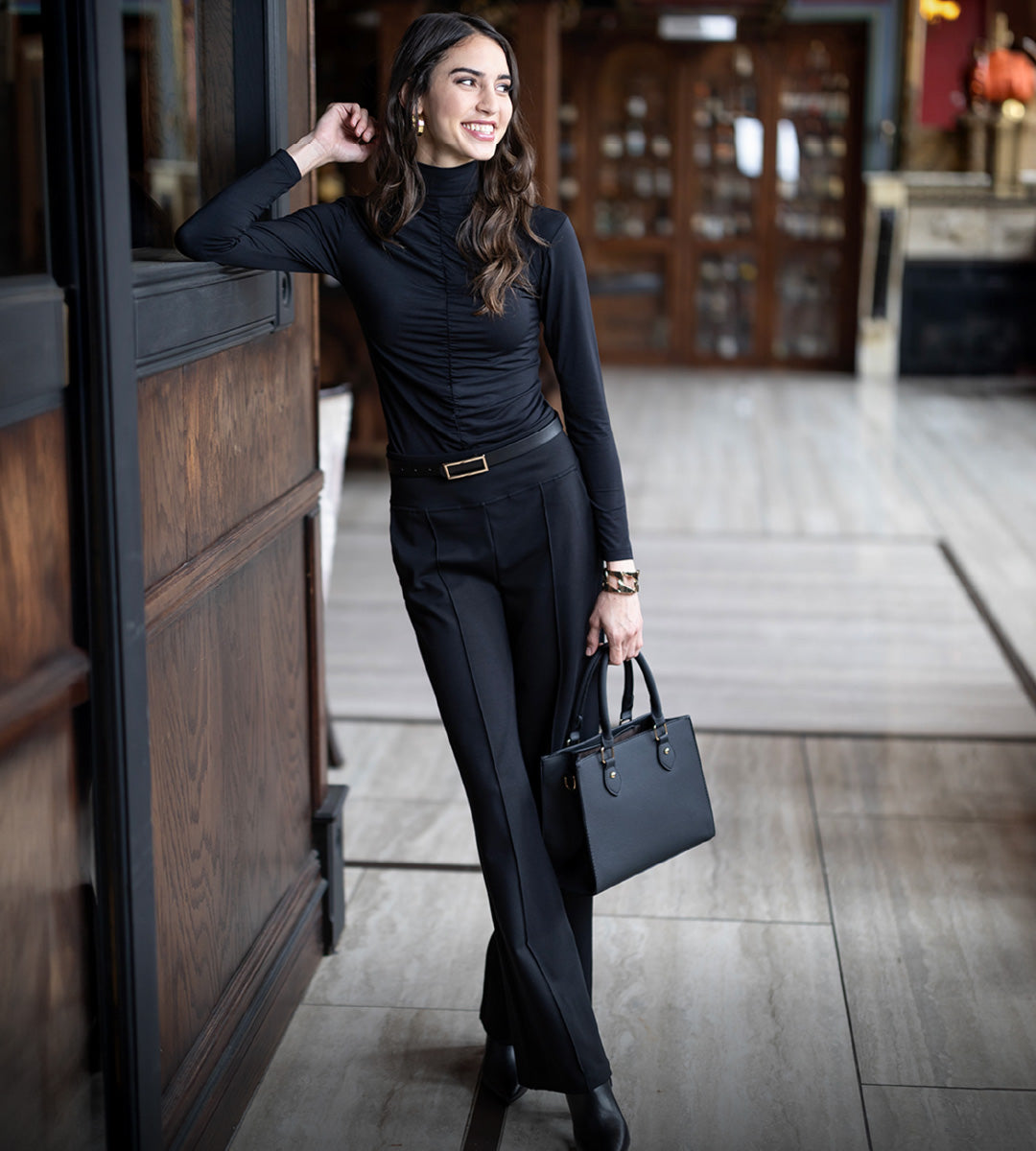 Woman in a black ruched top and black high waisted pintuck pants holding a black handbag in an indoor setting.