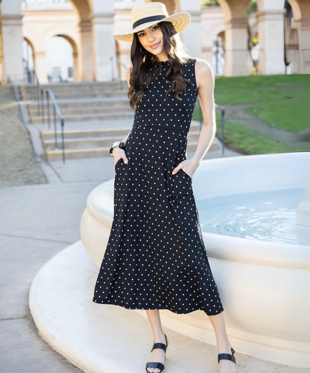 Woman wearing a black sleeveless polka dot maxi dress with pockets, accessorized with a wide-brim straw hat, posing outdoors near a fountain.