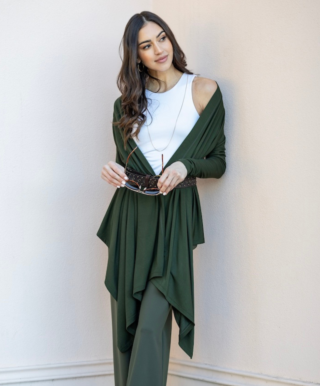 A woman wearing a white tank top and green cardigan, with a belt and green pants, standing near a wall.