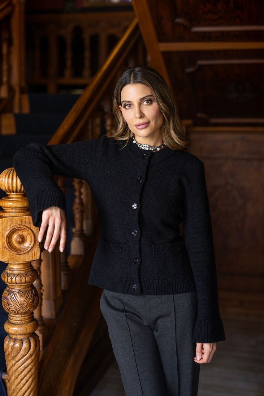 Woman in a black buttoned up cardigan and gray pants standing on wooden stairs.