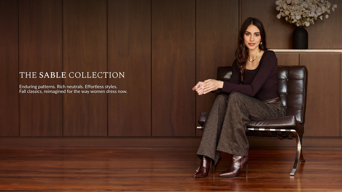 Woman sitting on leather seat wearing brown herringbone pants and a brown scoop neck long sleeve in front of a wood panel wall