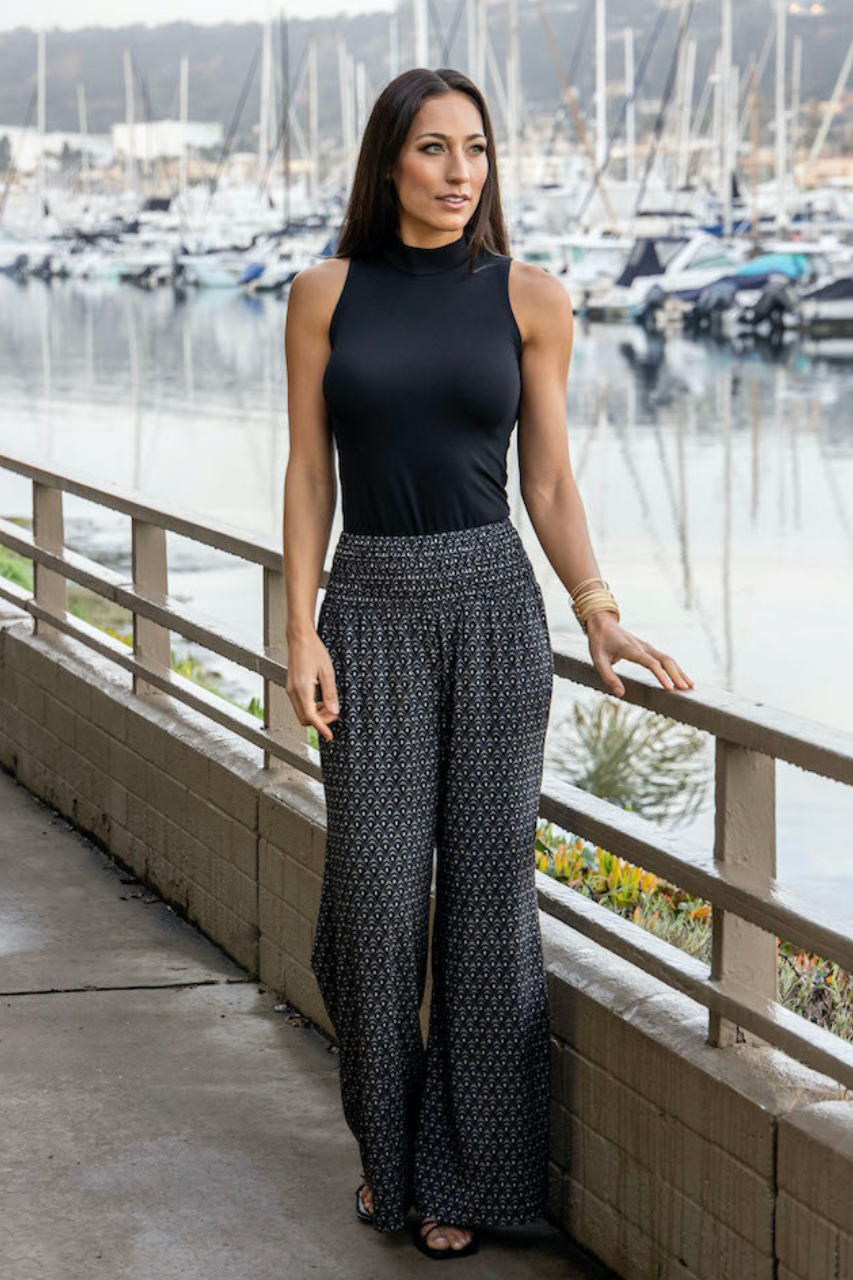 Woman by the water wearing a black top paired with the Portofino Travel Pant in woodblock print, creating a stylish and relaxed look perfect for a scenic day out.