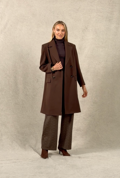 Woman walking onto a studio background wearing a brown coat, brown herringbone pants, and brown boots. She poses for a second, spins, and walks off