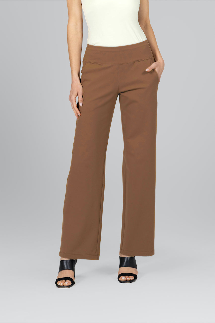 A woman wearing dark khaki traveler pants with a white top, standing against a grey background.
