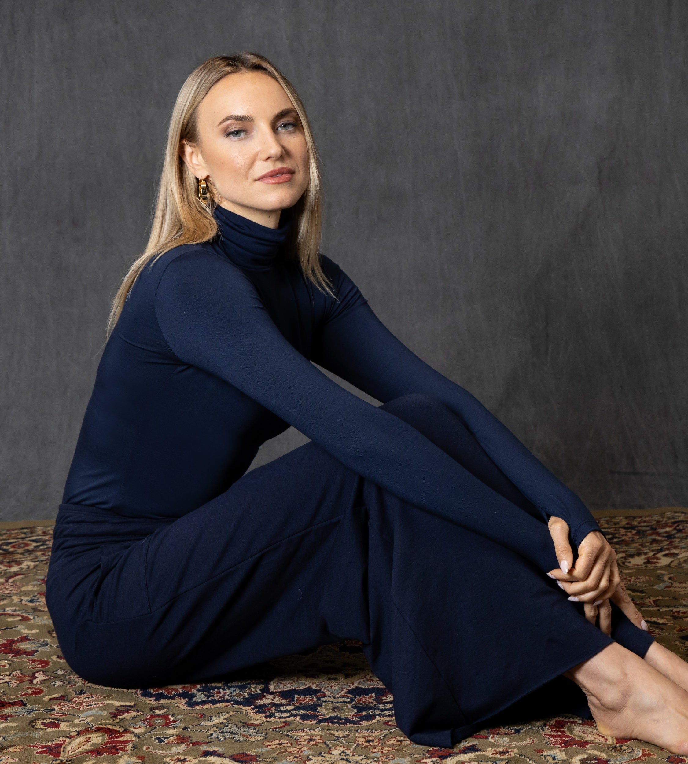 Woman in a navy blue outfit and cozy navy blue pants sitting on a patterned rug against a dark gray background