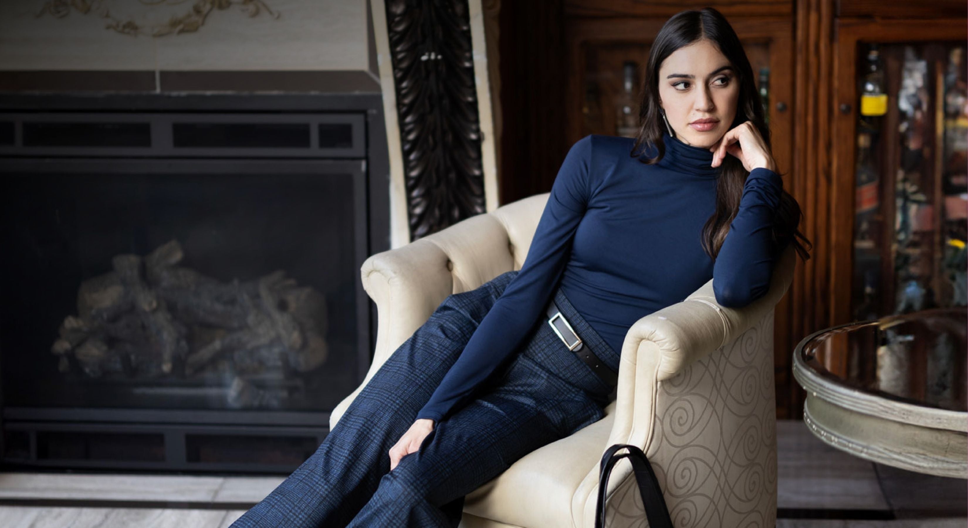 Woman in a blue turtleneck and plaid pants outfit sitting on a white chair in a room with a fireplace and wooden furniture.